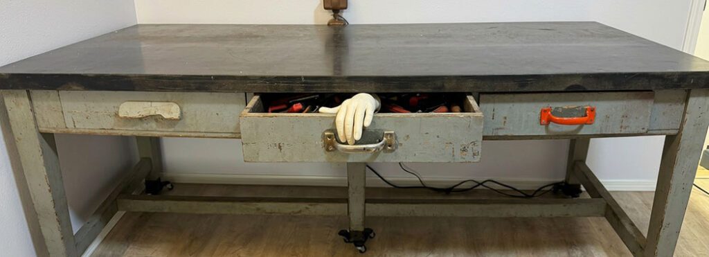 Photographic-realistic illustration of a vintage industrial lab bench with weathered driftwood-gray drawer fronts and a deep slate-black scratched top, the central drawer pulled open with a bright orange-red utility handle mounted on the front and a pale cream prosthetic hand emerging from inside the drawer curled over the back of the handle, against soft dove-cream walls and warm honey-cream wood-plank flooring