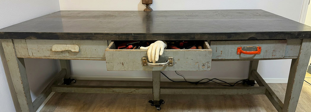 Photographic-realistic illustration of a vintage industrial lab bench with weathered driftwood-gray drawer fronts and a deep slate-black scratched top, the central drawer pulled open with a bright orange-red utility handle mounted on the front and a pale cream prosthetic hand emerging from inside the drawer curled over the back of the handle, against soft dove-cream walls and warm honey-cream wood-plank flooring